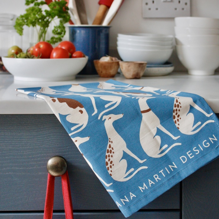 A photograph of a tea towel hanging off a kitchen side. The tea towel is blue and decorated with white and brown and whippets. White bowls decorate the side, with a dish of tomatoes. 