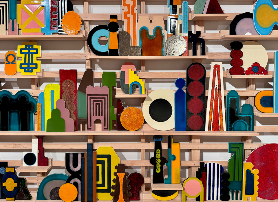 A series of glazed ceramic geometric shapes in bold, bright colours arranged on a wooden shelving structure.