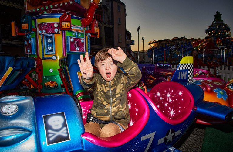 A photograph of a young boy sat on a fairground ride. He is sat in an airplane with a happy expression on his face, raising both arms up.
