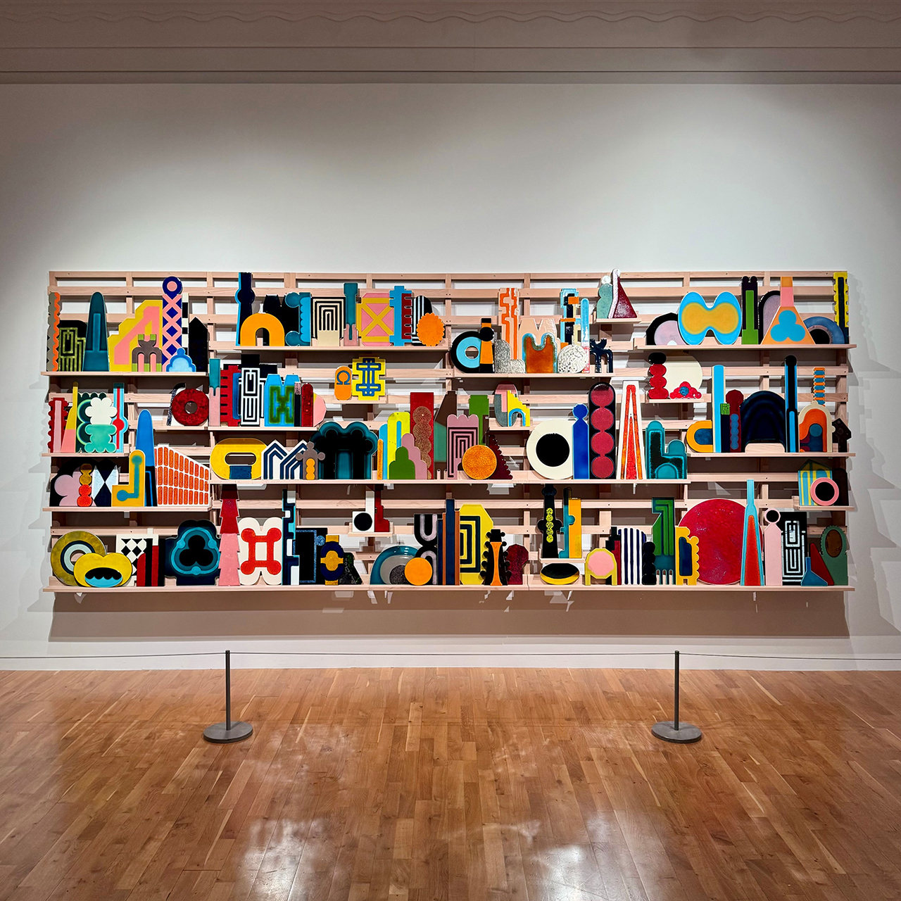 A photograph of a large, brightly coloured artwork in an art gallery. The work is made up of many small geometric shapes arranged on a wooden structure with three narrow shelves.  