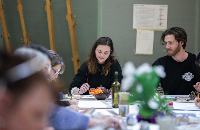 A photograph of a man and a woman sat at a table full of art materials and objects to draw. The woman is holding a paintbrush.