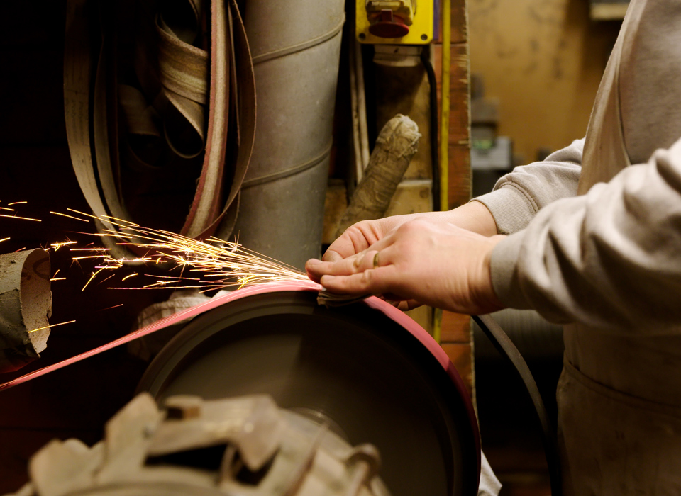 A torso and hands of a person using a grinding wheel and creating sparks. In the background there is a workshop with tubing, switches and machinery.