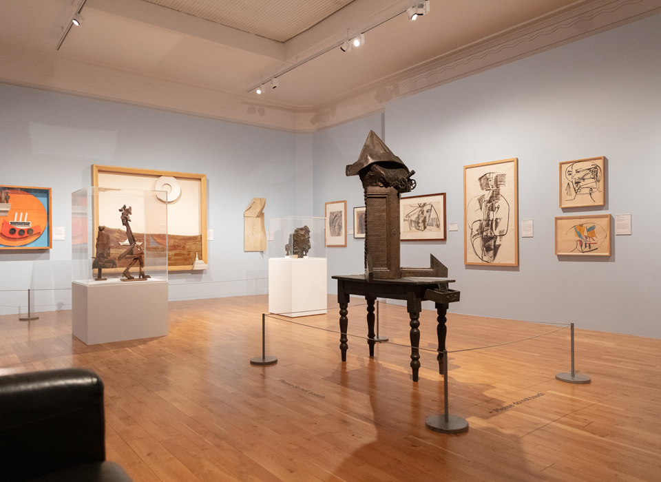 An exhibition space with light blue wall showing predominantly monochrome drawings on the walls, In the foreground is a dark, freestanding abstract sculpture. Behind it are two smaller sculptures sat on white plinths and surrounded by Perspex cases.