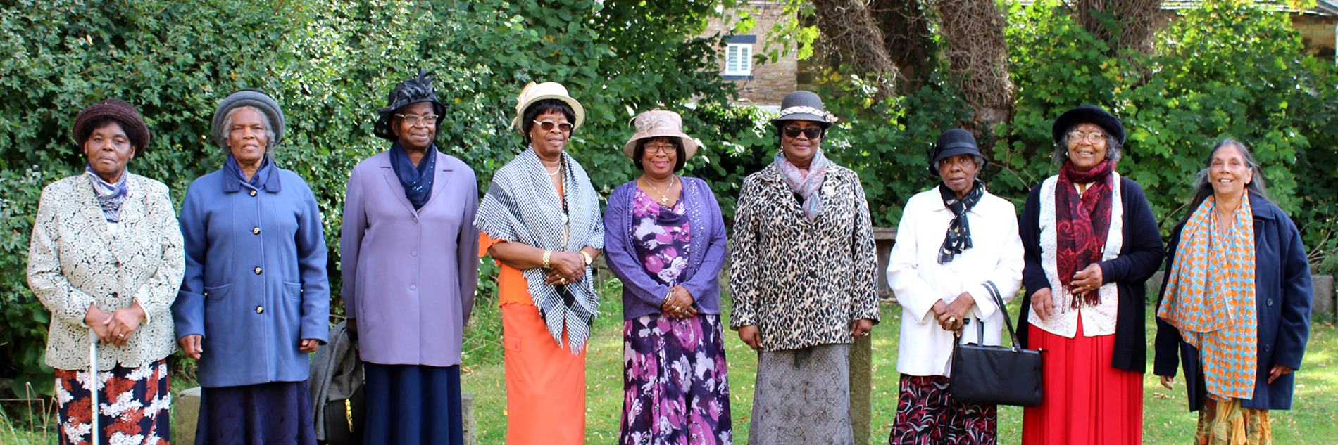 A photograph of the Black Ladies Group. There are eight people standing in a line, some are wearing hats. They are outside with trees behind them.