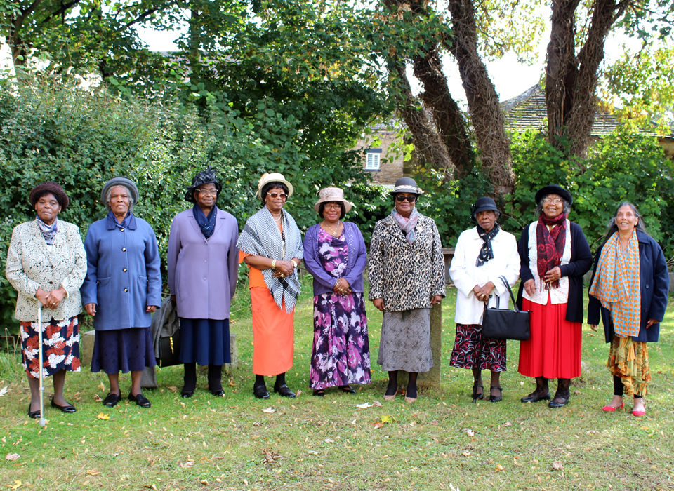 A photograph of the Black Ladies Group. There are eight people standing in a line, some are wearing hats. They are outside with trees behind them.