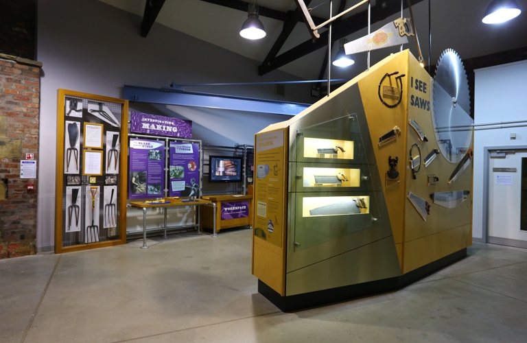 A photograph of displays in a museum gallery. There are garden forks, saws, a large saw and a large circular saw on display.   