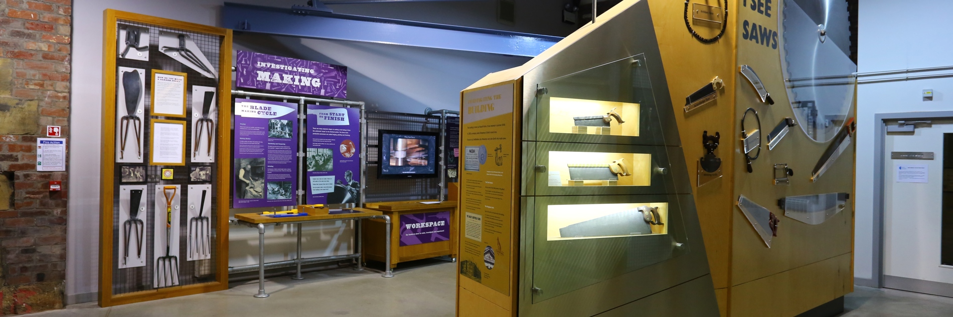 A photograph of displays in a museum gallery. There are garden forks, saws, a large saw and a large circular saw on display.   