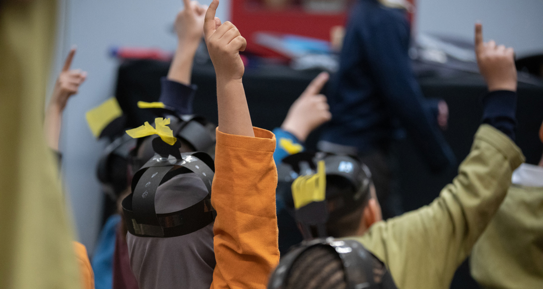 A view from behind of several primary school aged children with raised hands. They are dressed as Anglo Saxons and wearing brightly coloured tunics and paper helmets. 