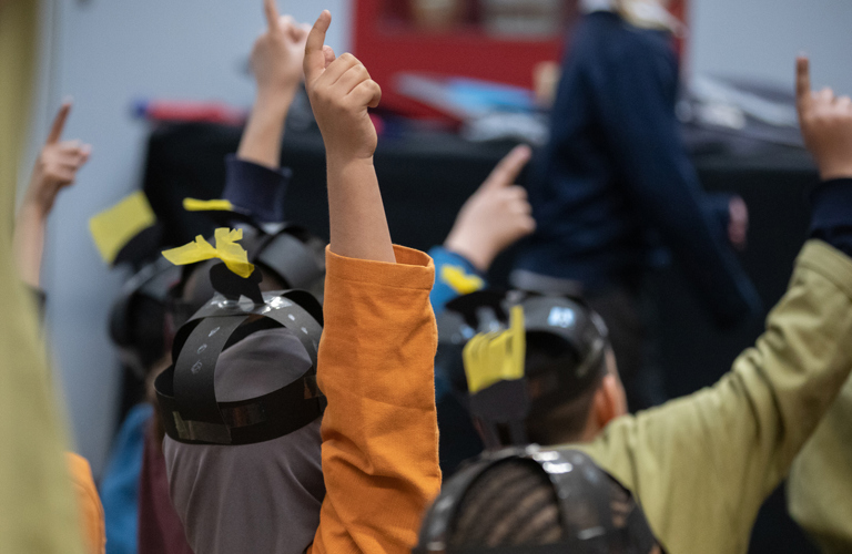 A view from behind of several primary school aged children with raised hands. They are dressed as Anglo Saxons and wearing brightly coloured tunics and paper helmets.