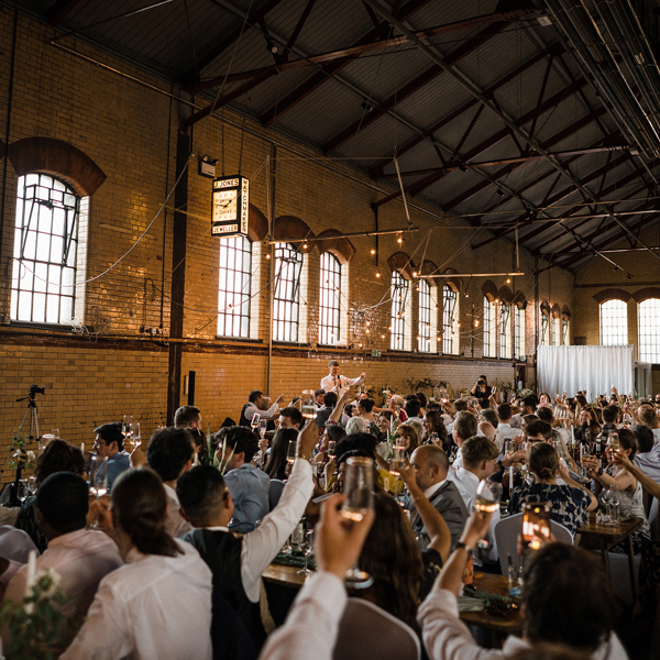 Guest raise their glasses for a toast in a large room of an industrial building decorated with fairly lights.