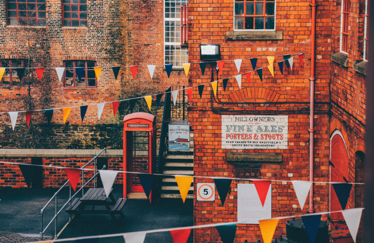 A photograph of industrial buildings, a phone box and a bench. Bunting is hung between the buildings. A sign on the building says "The Millowners Arms" and on the side of the wall there is a large painted sign which says "Millowners fine ales, porters and stouts". 