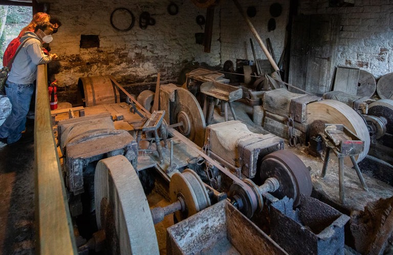 An old workshop with three grindstones. Leaning against the walls are piles of wood, tools and stones of different shapes and sizes.