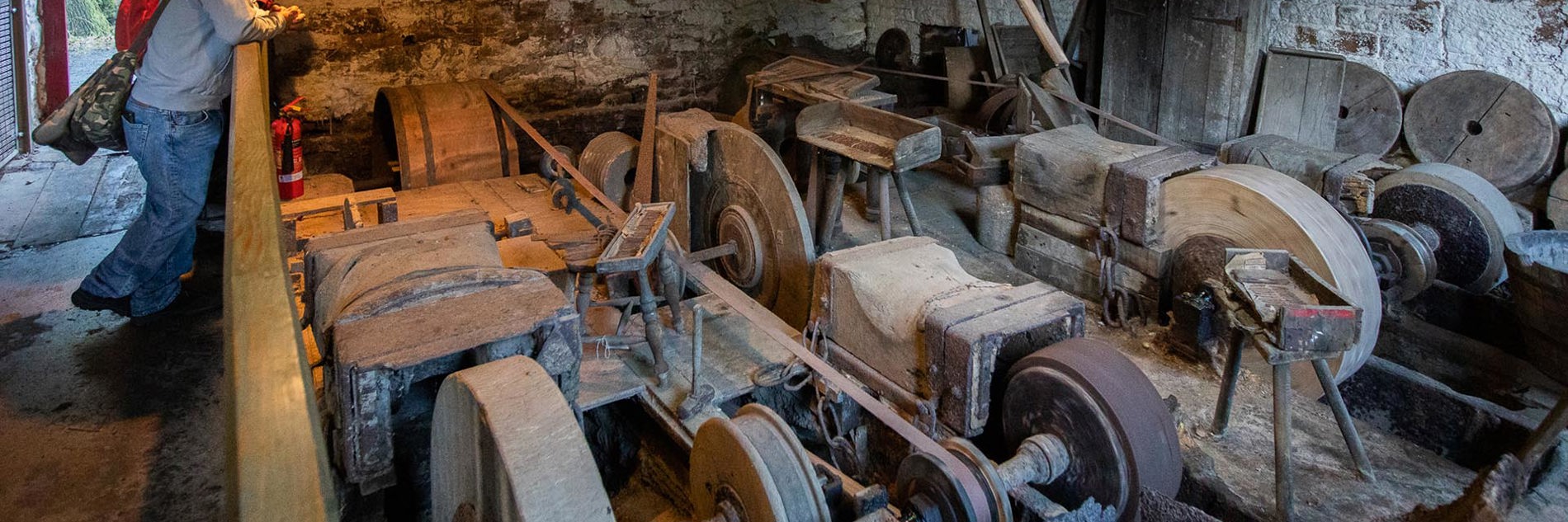 An old workshop with three grindstones. Leaning against the walls are piles of wood, tools and stones of different shapes and sizes.