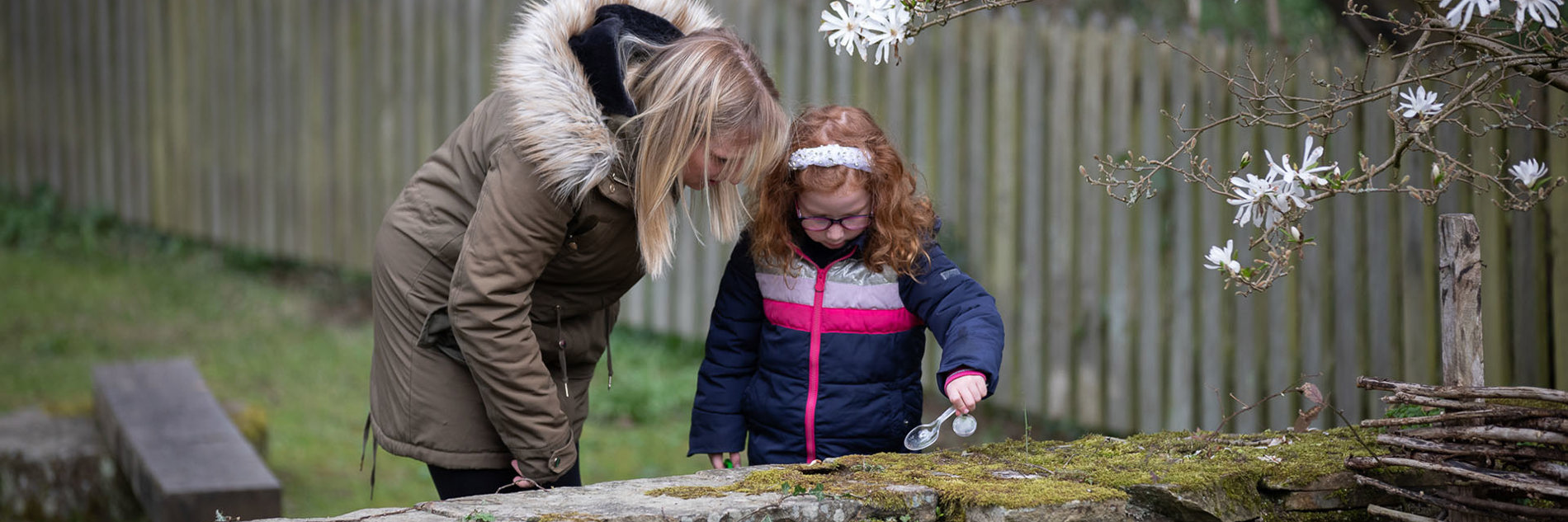 An adult and child looking at a moss covered wall. The child is using a spoon to collect a sample.