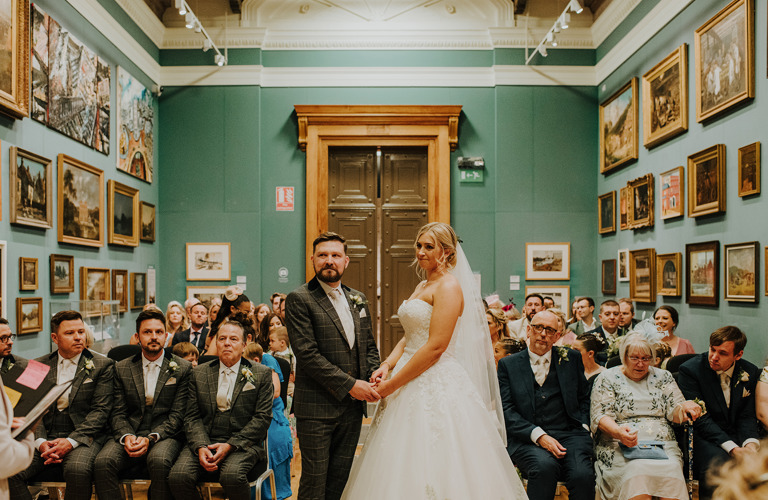 A groom in a suit and a bride in wedding dress stand in a gallery with with pictures in frames covering the walls on either side. behind the couple, wedding guests are seated either side of an aisle.