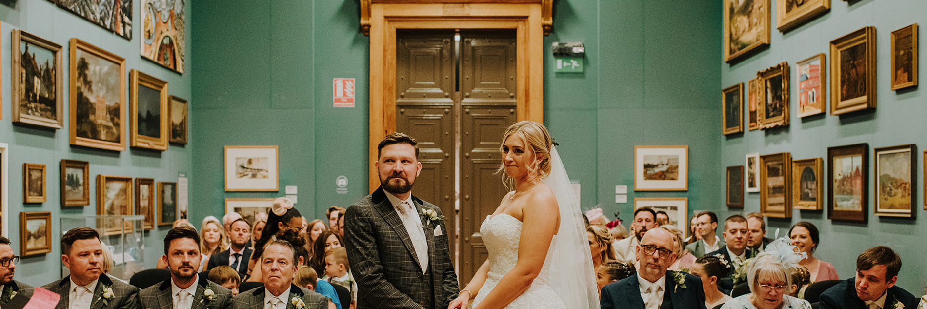 A groom in a suit and a bride in wedding dress stand in a gallery with with pictures in frames covering the walls on either side. behind the couple, wedding guests are seated either side of an aisle.