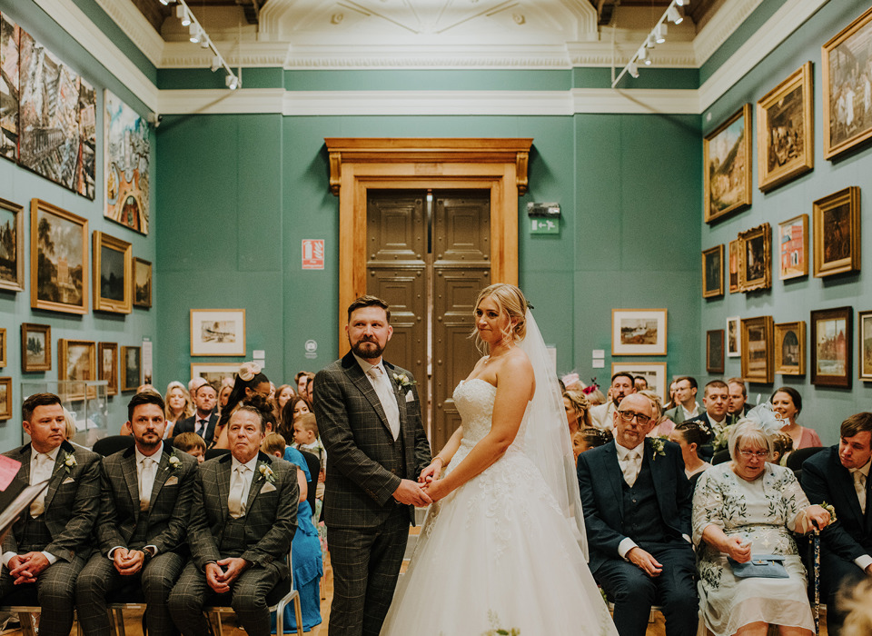 A groom in a suit and a bride in wedding dress stand in a gallery with with pictures in frames covering the walls on either side. behind the couple, wedding guests are seated either side of an aisle.