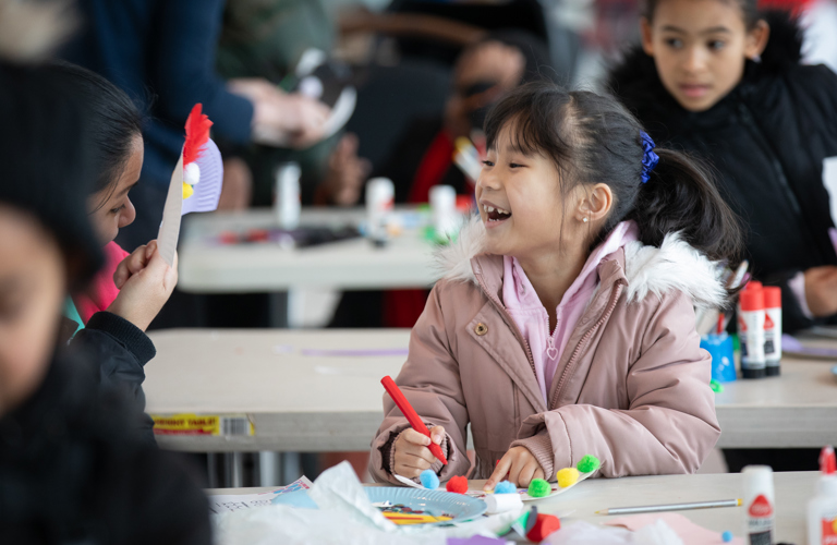 A child smiling and looking at an adult who is holding a decorated piece of paper. The child and adult are sitting a table with craft supplies, and the child is holding a pen. Other adults and children can be seen in the background.  