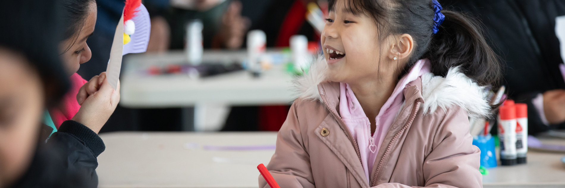 A child smiling and looking at an adult who is holding a decorated piece of paper. The child and adult are sitting a table with craft supplies, and the child is holding a pen. Other adults and children can be seen in the background.  