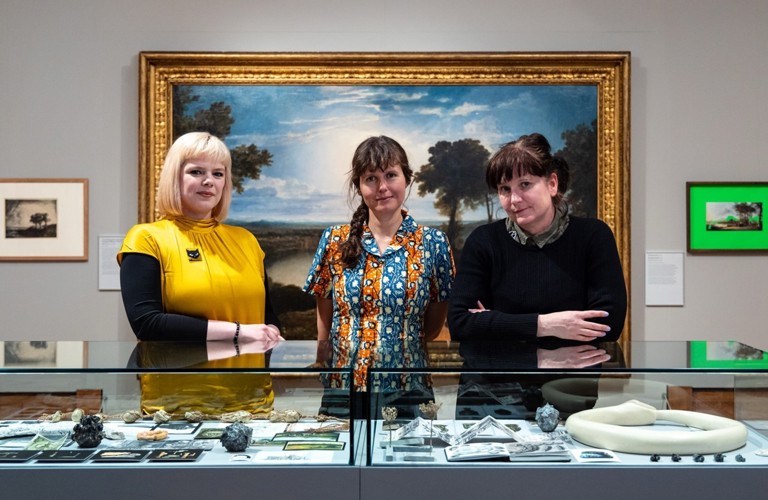 A photograph of three women behind a glass display cabinet of objects. On the wall behind them is a large landscape painting.