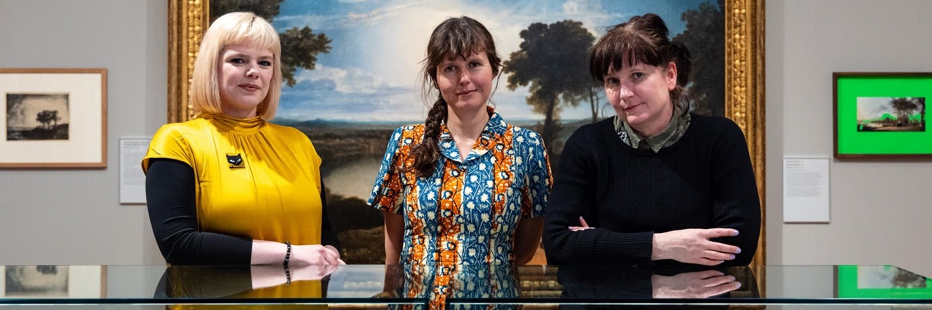 A photograph of three women behind a glass display cabinet of objects. On the wall behind them is a large landscape painting.