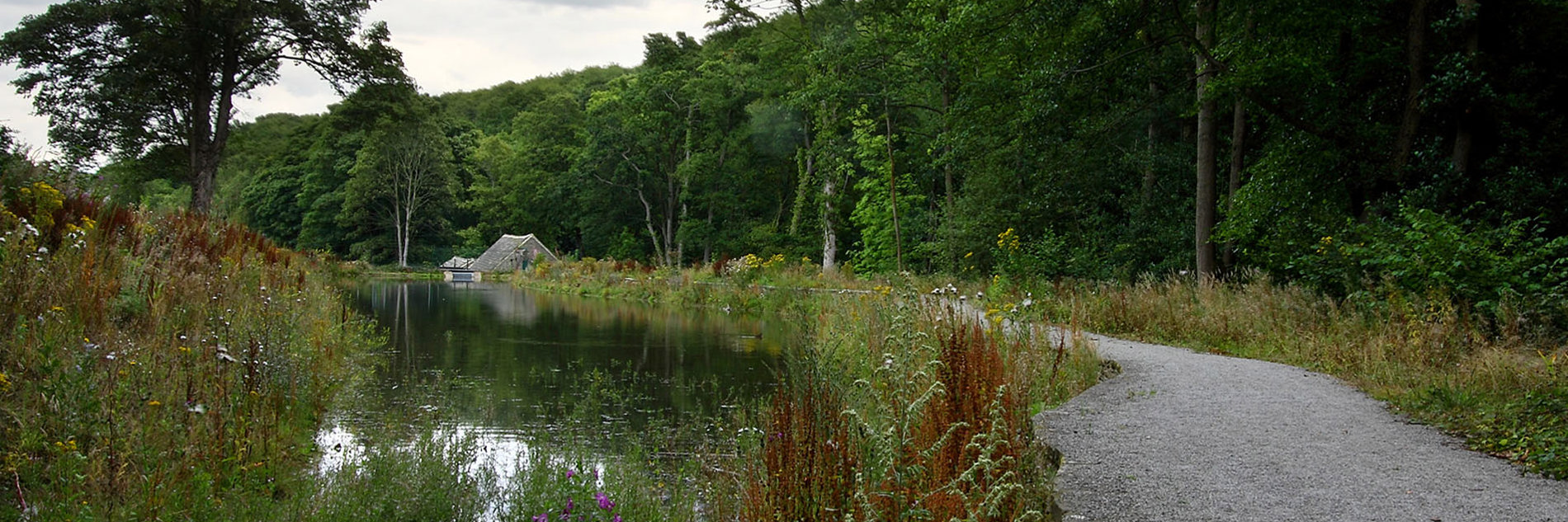 A body of water with a stone path that follows the edge of the water. The path leads to a small building which is surrounded by trees and vegetation. 