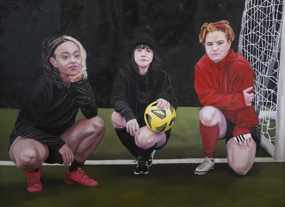 A painting of three teenage girls in football gear, all crouched next to the goal. The girl in the centre holds a yellow ball, all look directly out of the painting towards the viewer. 