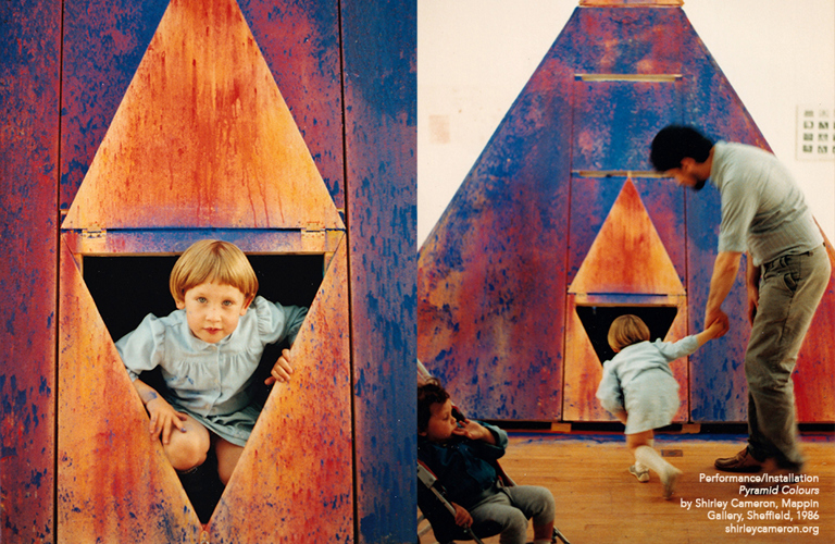 A photograph of a colourful art installation. L-R: a child is looking out from a triangle shaped window; an adult and child are looking into a triangle shaped window.  