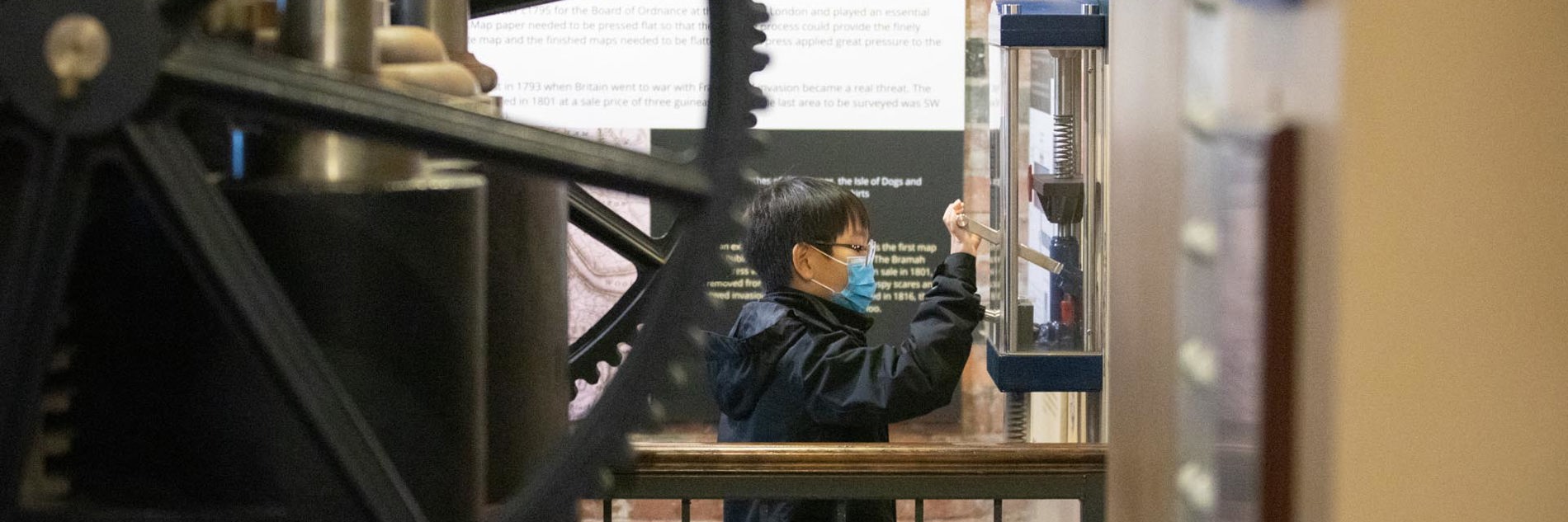 A child wearing a medical facemask operating an interactive scale model of a hydraulic press alongside the real thing.