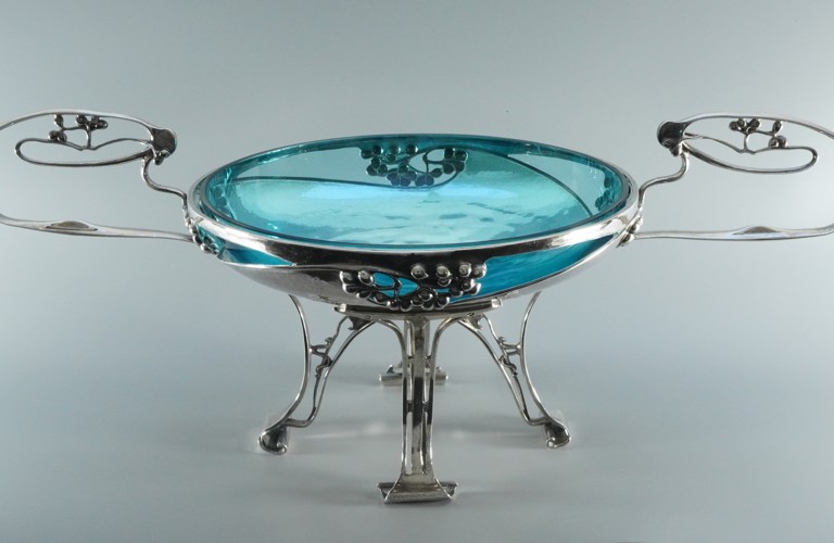 A photograph of a blue glass bowl, elevated on a silver, art deco base with four slender legs, and two curving handles on either side.