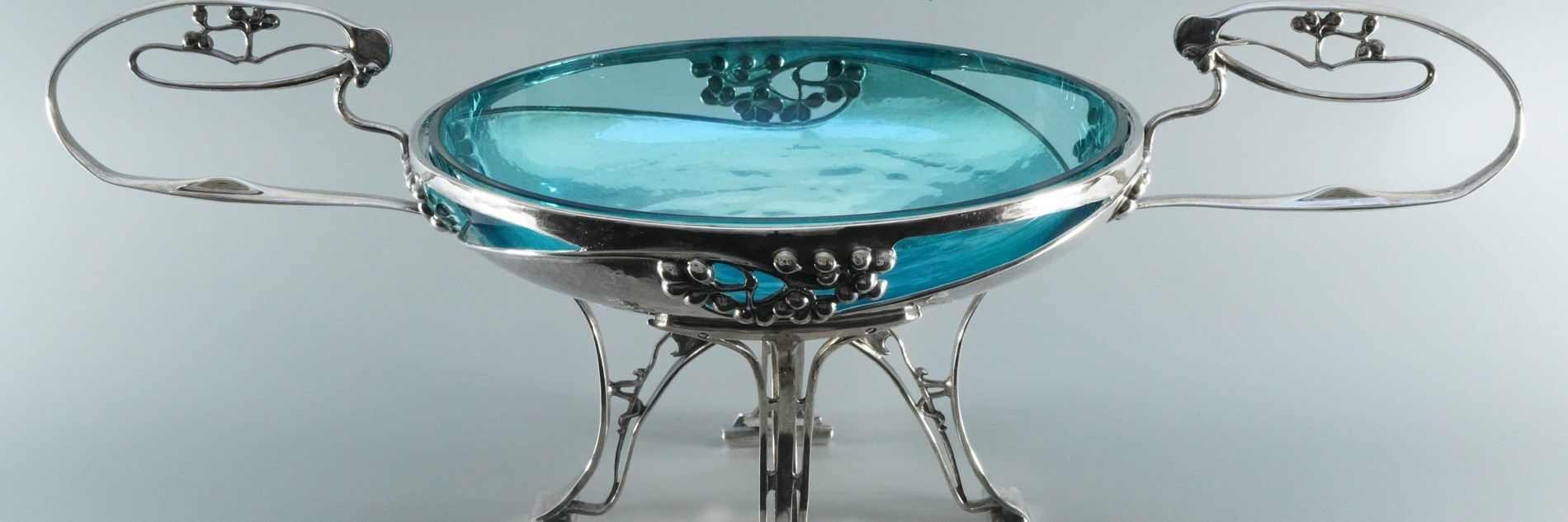 A photograph of a blue glass bowl, elevated on a silver, art deco base with four slender legs, and two curving handles on either side.