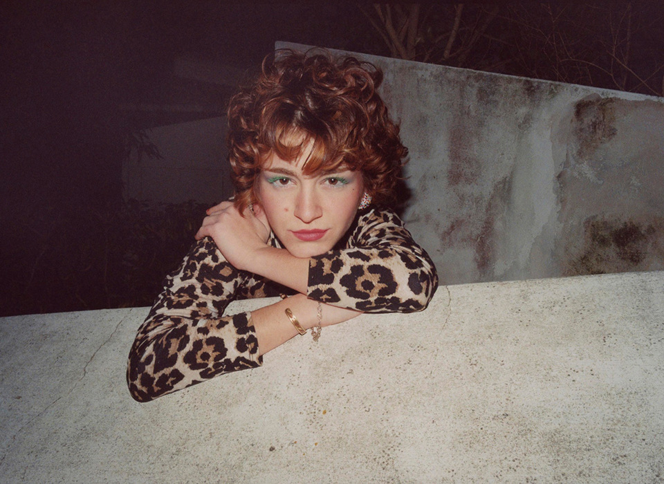 A woman wearing a leopard print dress leans over a wall, looking directly at the camera with a confident gaze. She has green mascara on her eyelashes, red curly hair and wears golden jewellery.