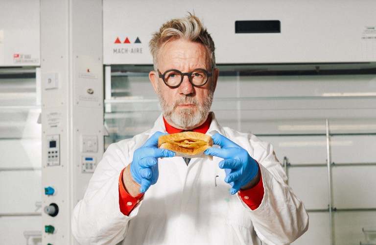A photograph of a man in a white lab coat, looking directly into the camera lens, holding up a crisp buttie for the viewer. He wears round black glasses and has grey hair and beard. He is stood in a lab environment with pristine white machinery behind him. He wears blue food safety gloves and a neutral expression.