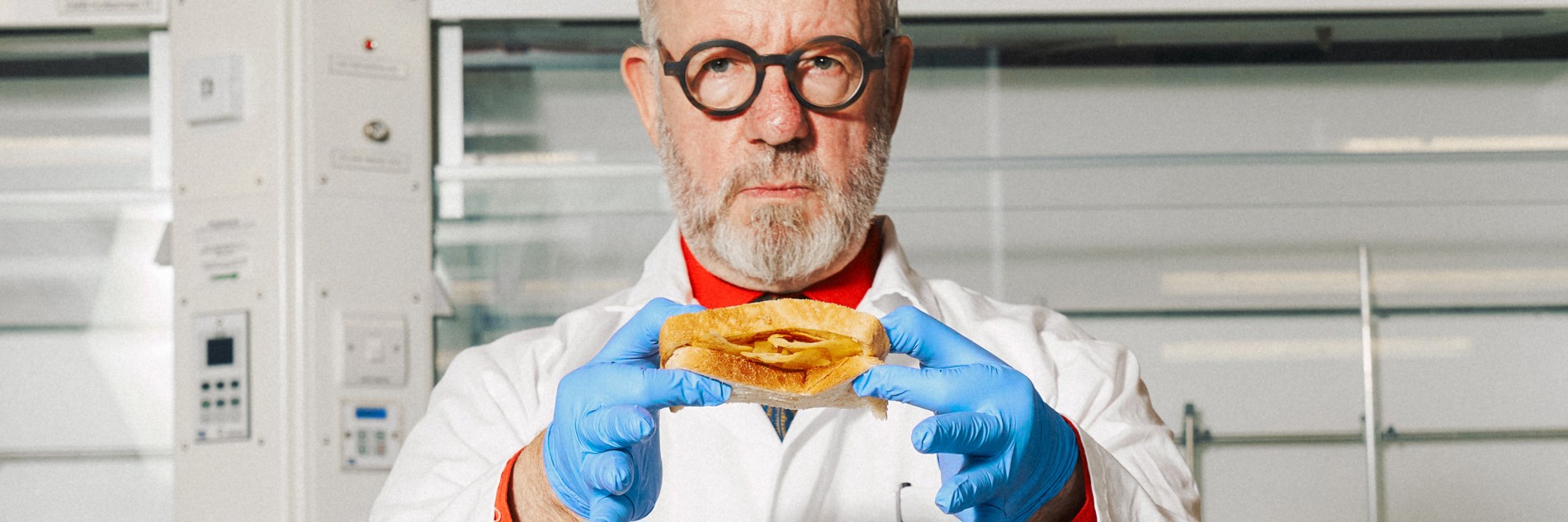 A photograph of a man in a white lab coat, looking directly into the camera lens, holding up a crisp buttie for the viewer. He wears round black glasses and has grey hair and beard. He is stood in a lab environment with pristine white machinery behind him. He wears blue food safety gloves and a neutral expression.
