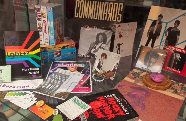 A series of objects from the Proud! display at Weston Park Museum. There is an LGBT+ handbook from 2018, a Queer as Folk boxset, Madge Woollard CD case, club night flyer, and various badges and leaflets.