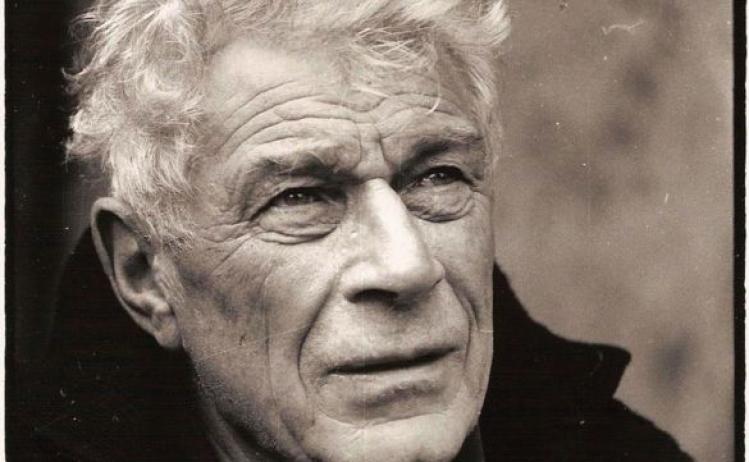 Black and white, close-crop portrait of John Berger, he's middle-aged with white hair, wearing a coat and his hair is being tousled in the wind, as if outdoors.