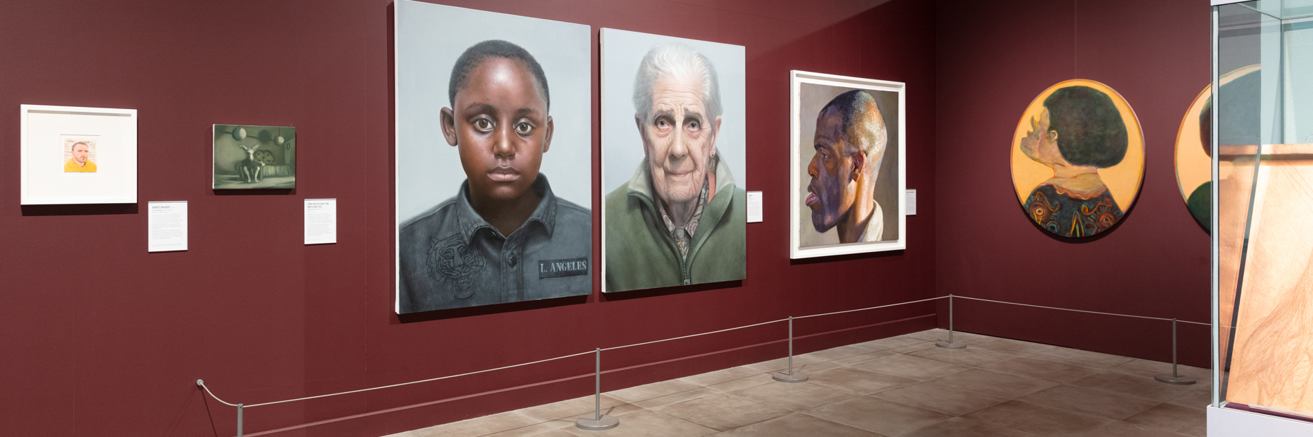 One corner of an exhibition in Millennium Gallery, with ox blood painted walls. There are seven portraits on the walls, varying in size.