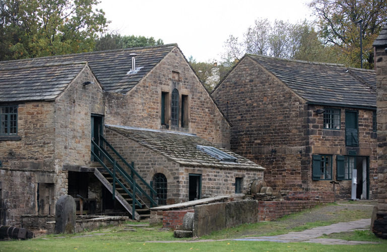 A collection of historic industrial stone buildings. They have large green windows and the building to the left has steps leading up to the second storey. There are trees behind.