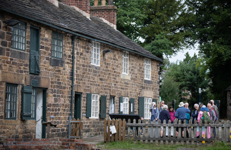 A group visitors gathered in front of a row of historic terraced stone cottage with trees in the background 