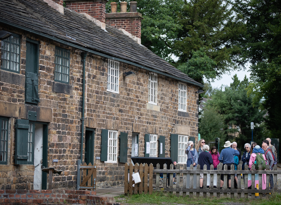 A group visitors gathered in front of a row of historic terraced stone cottage with trees in the background 