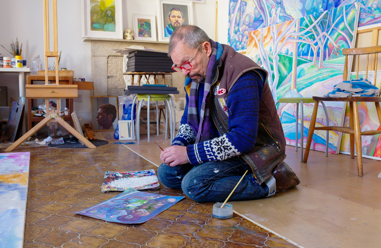 A photograph of an adult sitting on a wooden and lino floor and looking at an artwork. They have a paintbrush in their hand and a pot and paintbrush next to them. In the background there is an easel, a table with art supplies, and colourful artworks propped up again the wall and fireplace. 