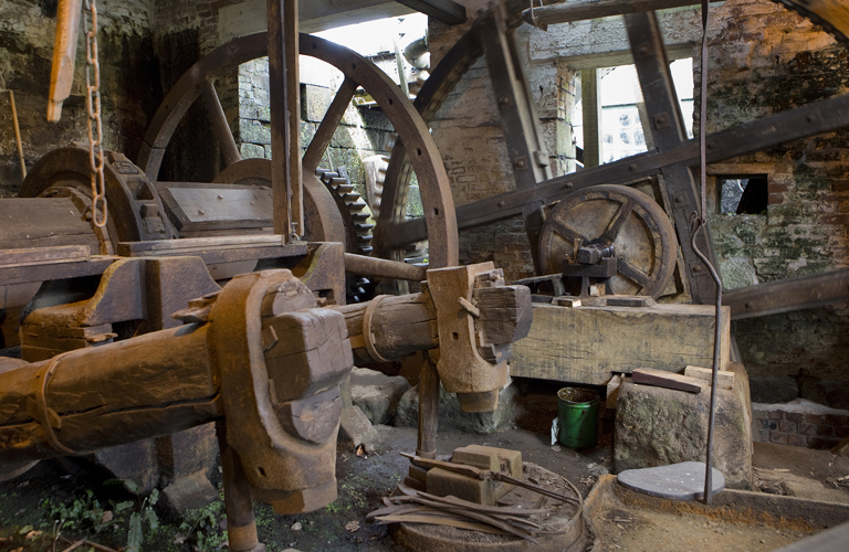 Two rusting tilt forge hammers mounted on huge old timber shafts with gearwheel drive mechanisms in the background.