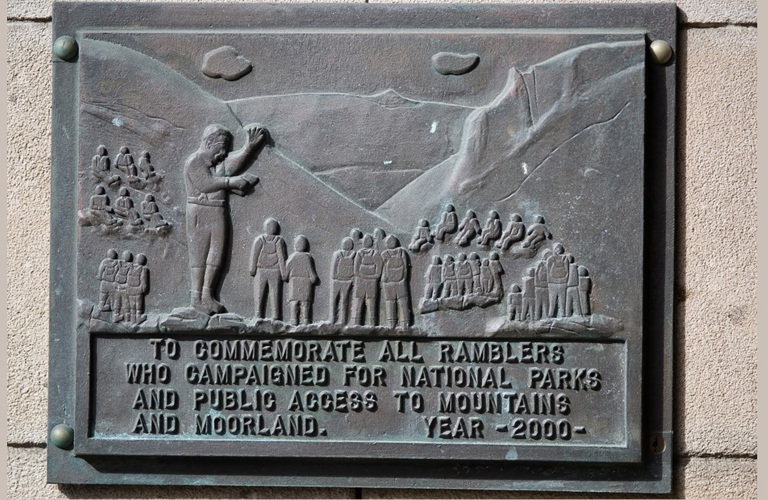 A commemorative stone relief plaque showing groups of ramblers walking and sitting, enjoying the countryside. There are hills in the background. The inscription reads "To commemorate all ramblers who campaigned for National Parks and public access to mountains and moorland. Year 2000".
