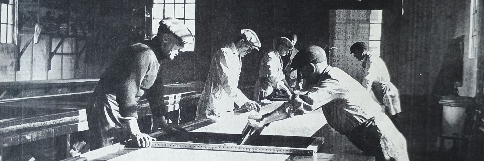 A black and white photo of workers in a screen printing factory around 1935. Six men are working around a long wooden table with metres of material stretched on top of it.