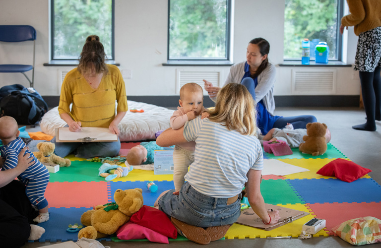 A group of babies and parents, sat on colourful mats, surrounded by baby toys and cushions. Some of the adults are drawing on large clipboards.