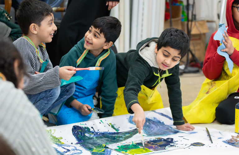 Children smile and chat as they kneel on teh floor and contribute to a large-scale artwork