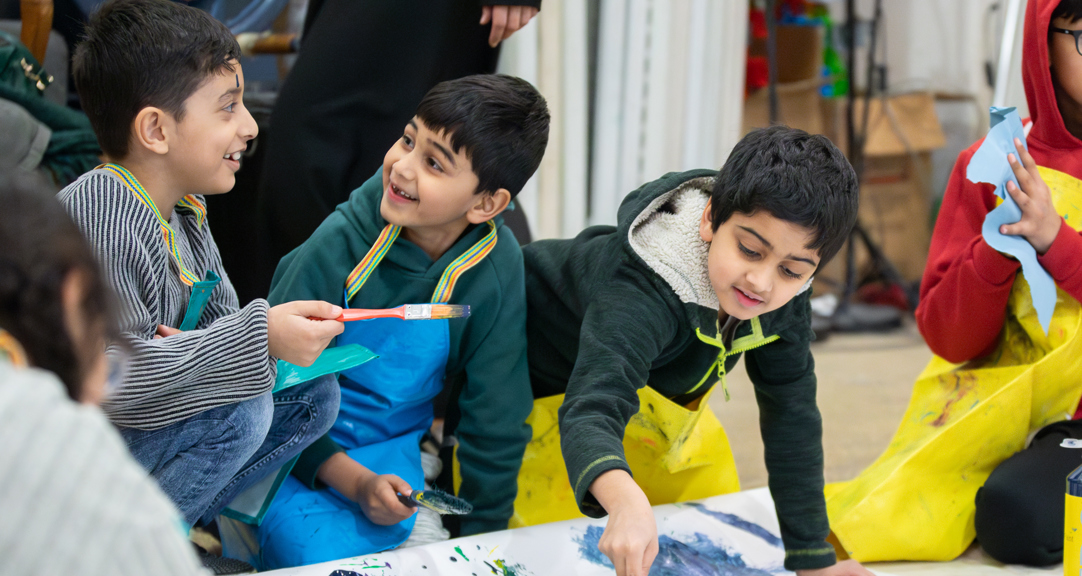 Children smile and chat as they kneel on teh floor and contribute to a large-scale artwork
