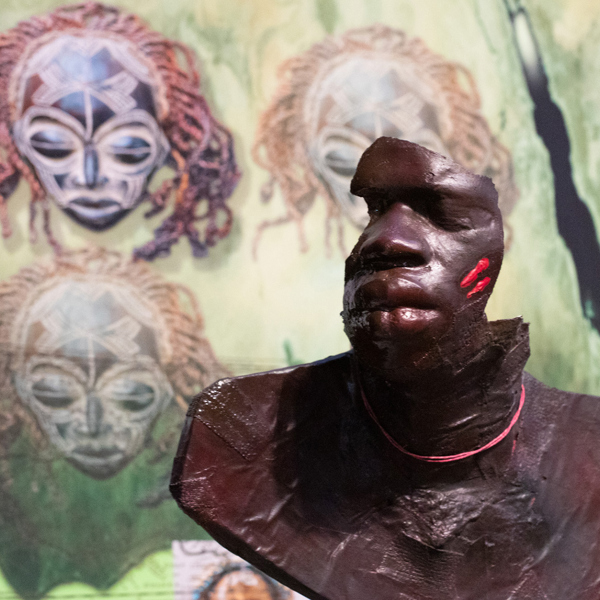 A detail photograph of Keith Piper's The Seven Rages Of Man. A broken bust of an black man is displayed in front of a graphic of tribal masks. 