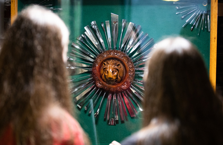 The back of two people's head as they look at a wooden lion carving surrounded by metal tools arranged around it like sun rays.