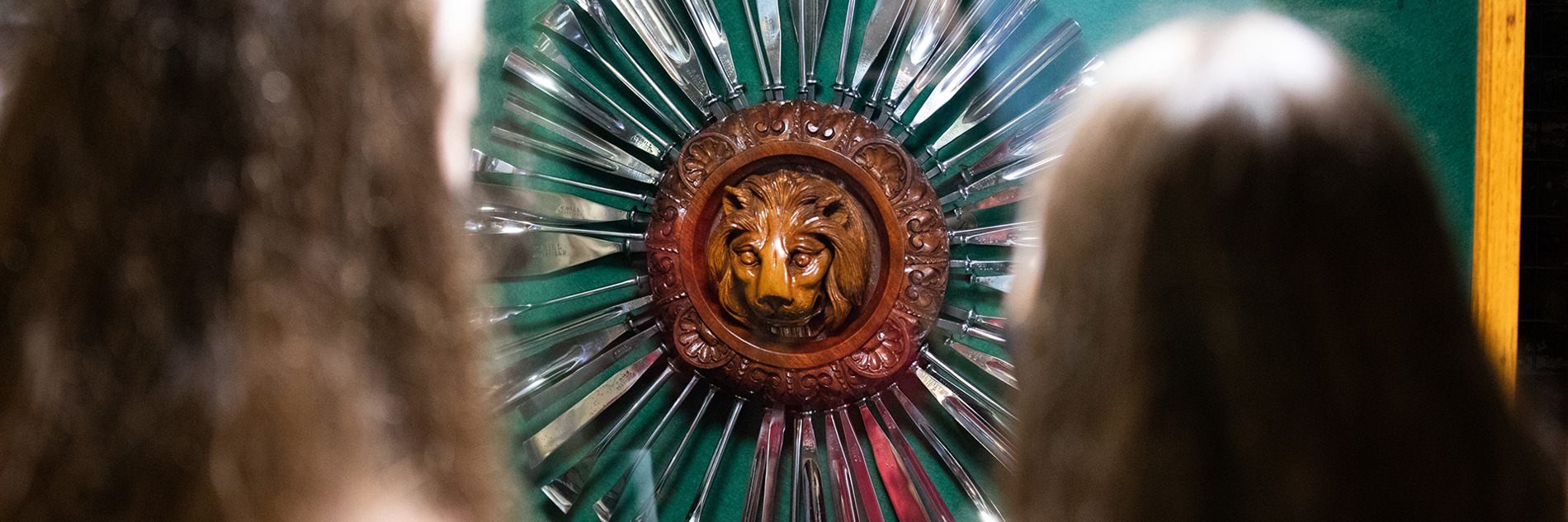 The back of two people's head as they look at a wooden lion carving surrounded by metal tools arranged around it like sun rays.